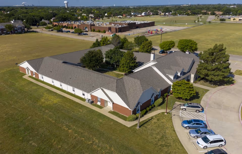 Aerial view of a single-story senior living facility building with a central courtyard filled with trees. The building has a gray roof and red brick and white exterior walls. There is a parking lot with several cars parked near the entrance. Surrounding the facility are open grassy fields and a few trees, with other buildings and a playground visible in the background.