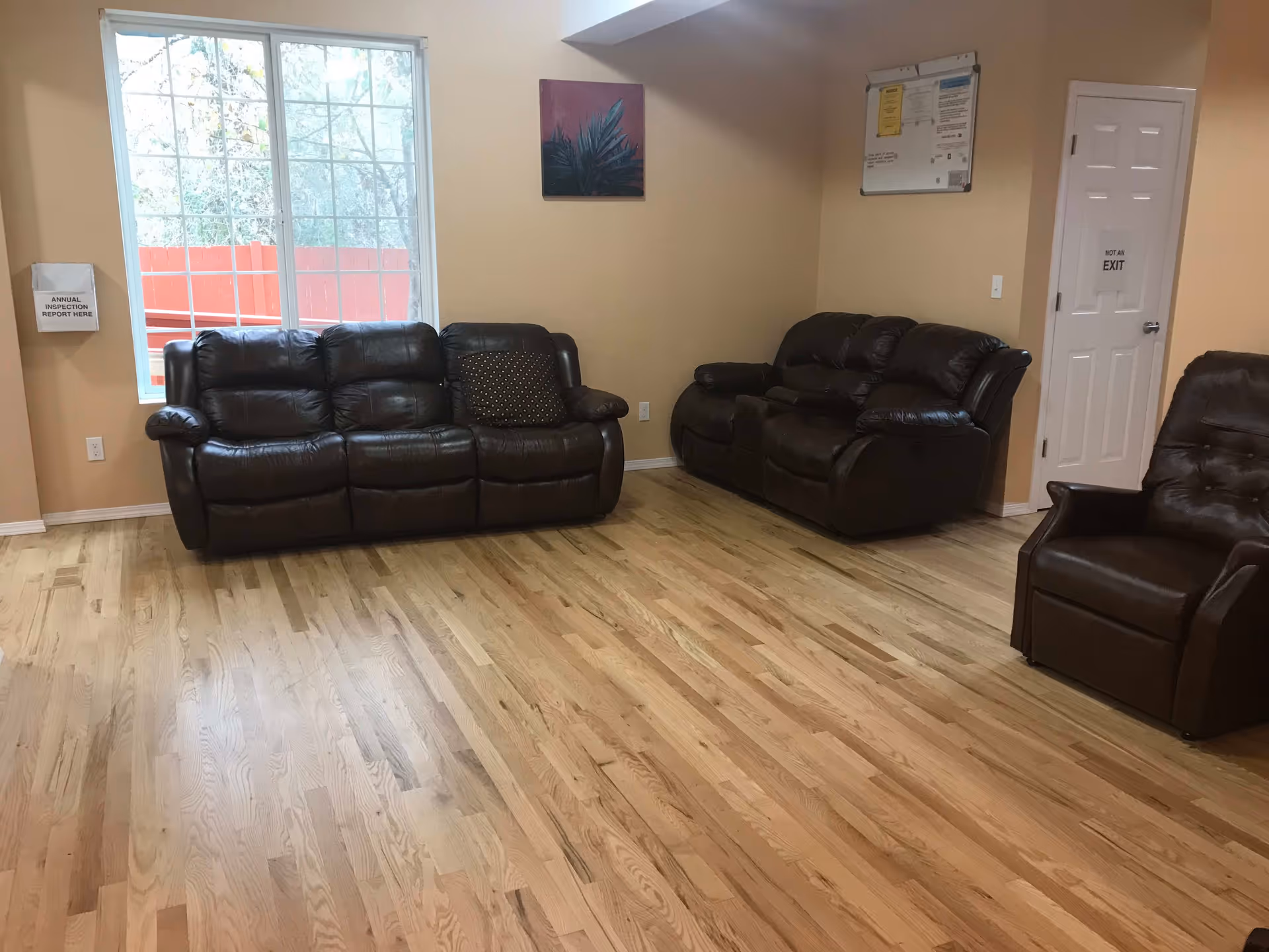 A living room with light wooden flooring, beige walls, and three dark brown leather seating pieces including two sofas and one armchair. A large window with white framing lets in natural light, and there is a small framed artwork on the wall. A white door with a sign that reads 'NOT AN EXIT' and a bulletin board are also visible on the right wall.