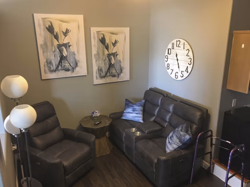 A cozy living room area with a dark leather recliner chair and matching loveseat with two blue pillows. A small wooden side table with a remote and a small decorative item sits between the chairs. Two abstract floral paintings hang on the wall above the recliner, and a large round wall clock is mounted above the loveseat. A floor lamp with three white globe lights stands next to the recliner. A purple walker is partially visible near the loveseat.