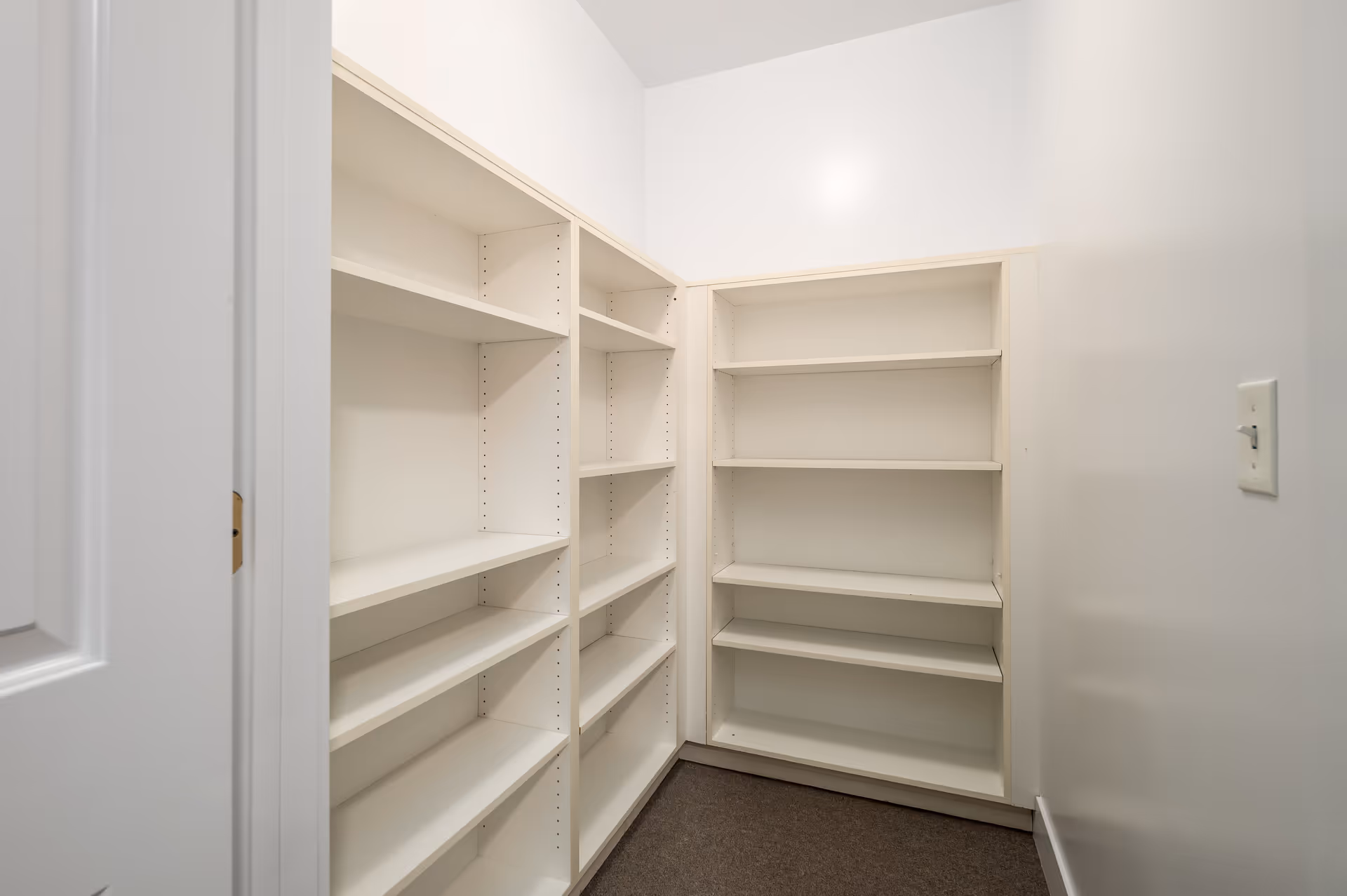A small empty storage room with white built-in shelves on two walls and a light switch on the right wall. The room has white walls and a carpeted floor.