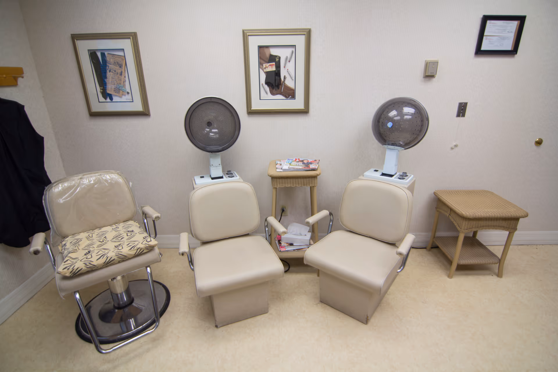 Interior view of a small salon area with two beige salon chairs under hair dryers, a third chair with a cushion, two wicker side tables with magazines and tissues, and framed pictures on the wall.