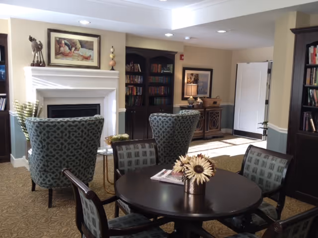 A cozy senior living common area with a round dark wooden table surrounded by four patterned chairs. Behind the table are two upholstered armchairs facing a white fireplace with a framed painting above it. To the right, there is a dark wooden bookshelf filled with books and a sideboard with a lamp and decorative items. The room has beige walls with white trim and recessed lighting in the ceiling.