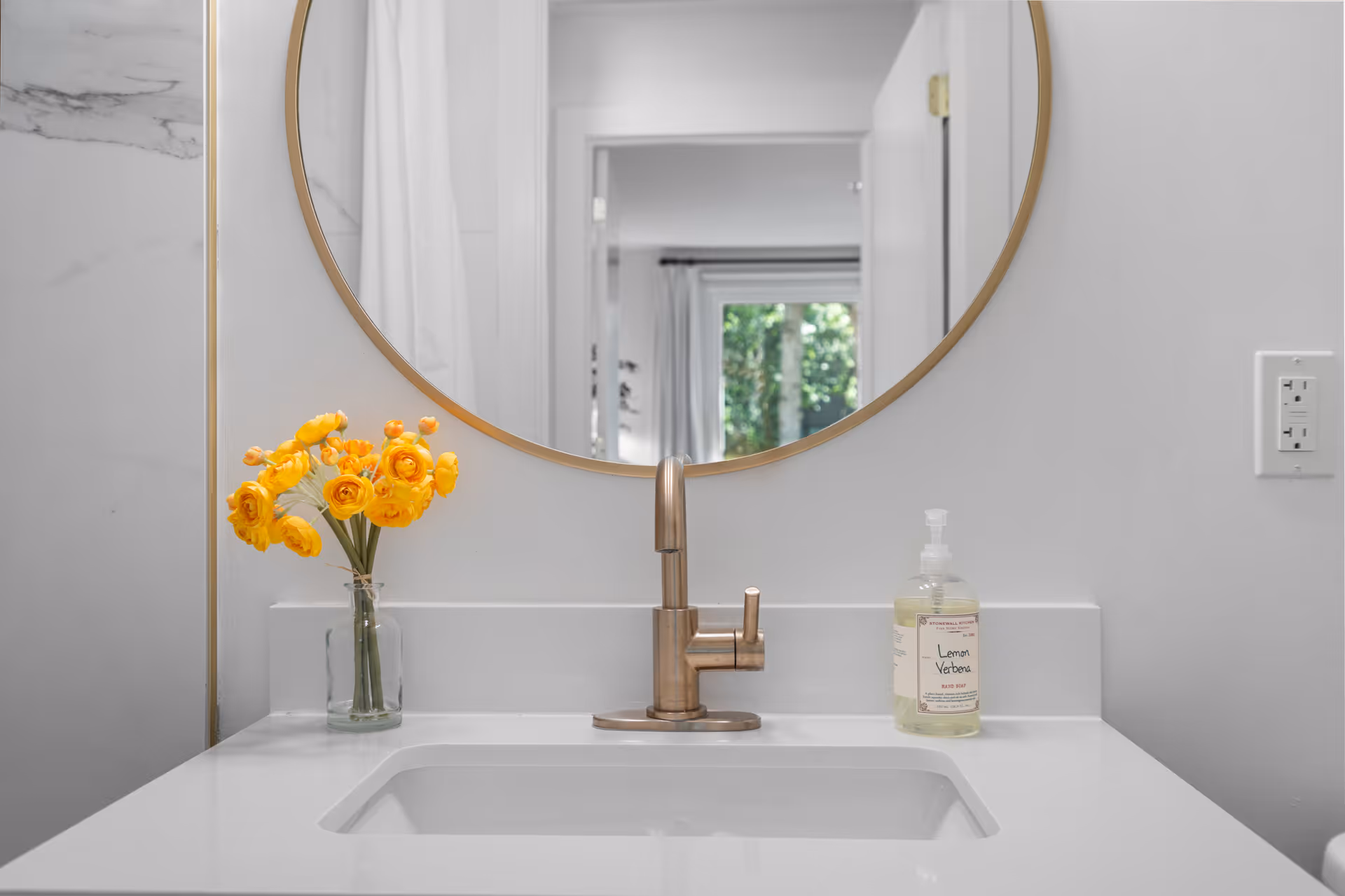 A modern bathroom sink with a gold faucet, a round mirror with a gold frame, a clear glass vase with yellow flowers on the left, and a bottle of lemon verbena hand soap on the right. The reflection in the mirror shows a doorway and a window with greenery outside.