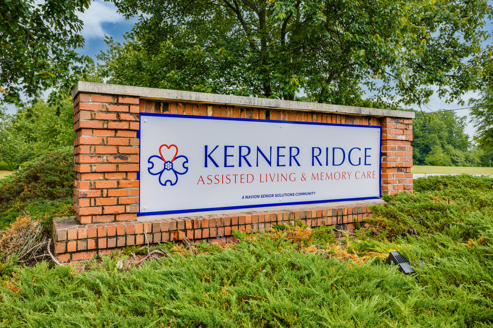 A brick sign for Kerner Ridge Assisted Living & Memory Care surrounded by green shrubs and trees under a partly cloudy sky.