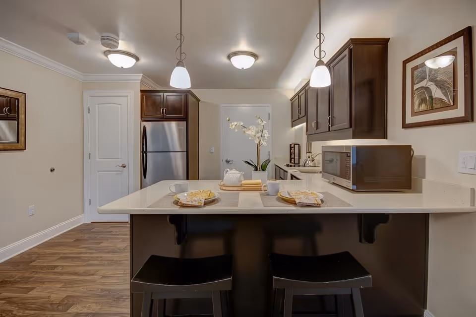Modern kitchen with dark wood cabinets, stainless steel refrigerator, microwave on the counter, and a kitchen island with two black stools. The island is set with plates, cups, napkins, and a teapot with a flower centerpiece. The room has wooden flooring and neutral-colored walls with a framed picture on the right wall.