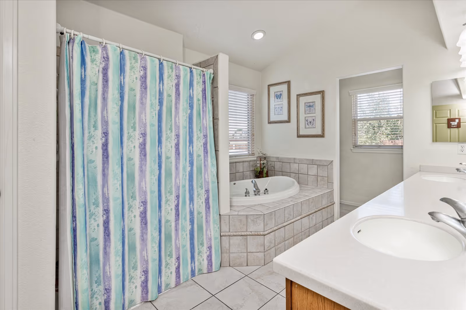 A bathroom with a tiled corner bathtub, a shower with a striped blue, green, and purple shower curtain, a double sink vanity with white countertops, two framed butterfly pictures on the wall, and two windows with blinds letting in natural light.