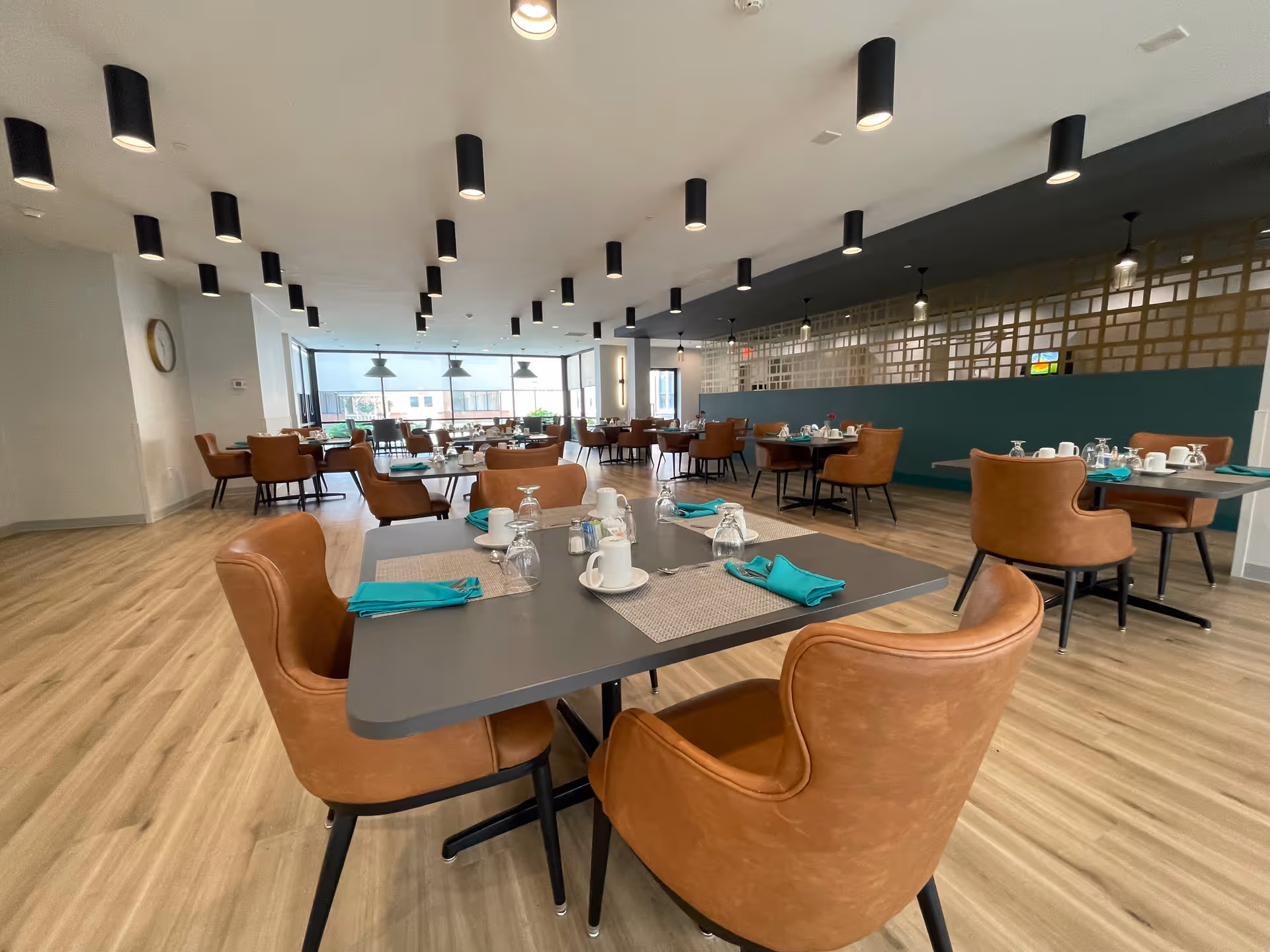Spacious modern dining room with multiple set tables, tan leather chairs, teal napkins, and overhead cylindrical lights.