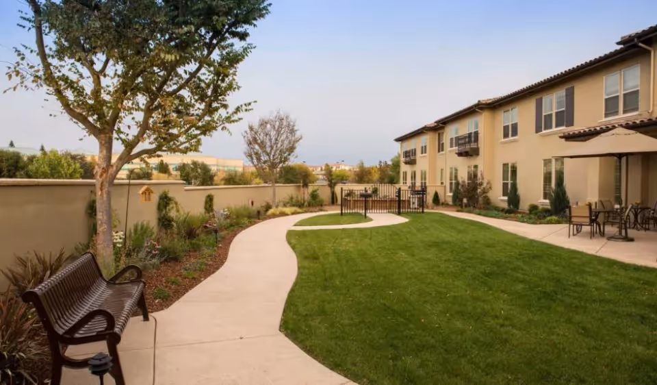 Outdoor courtyard area at Oakmont of Folsom featuring a curved concrete walkway, green lawn, trees, a bench, and a two-story beige building with windows and balconies. There are also patio tables with umbrellas on the right side.
