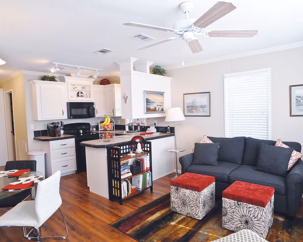 Open-plan living area and kitchen featuring a gray sofa with two patterned ottomans, a kitchen island with fruit, and white cabinets.