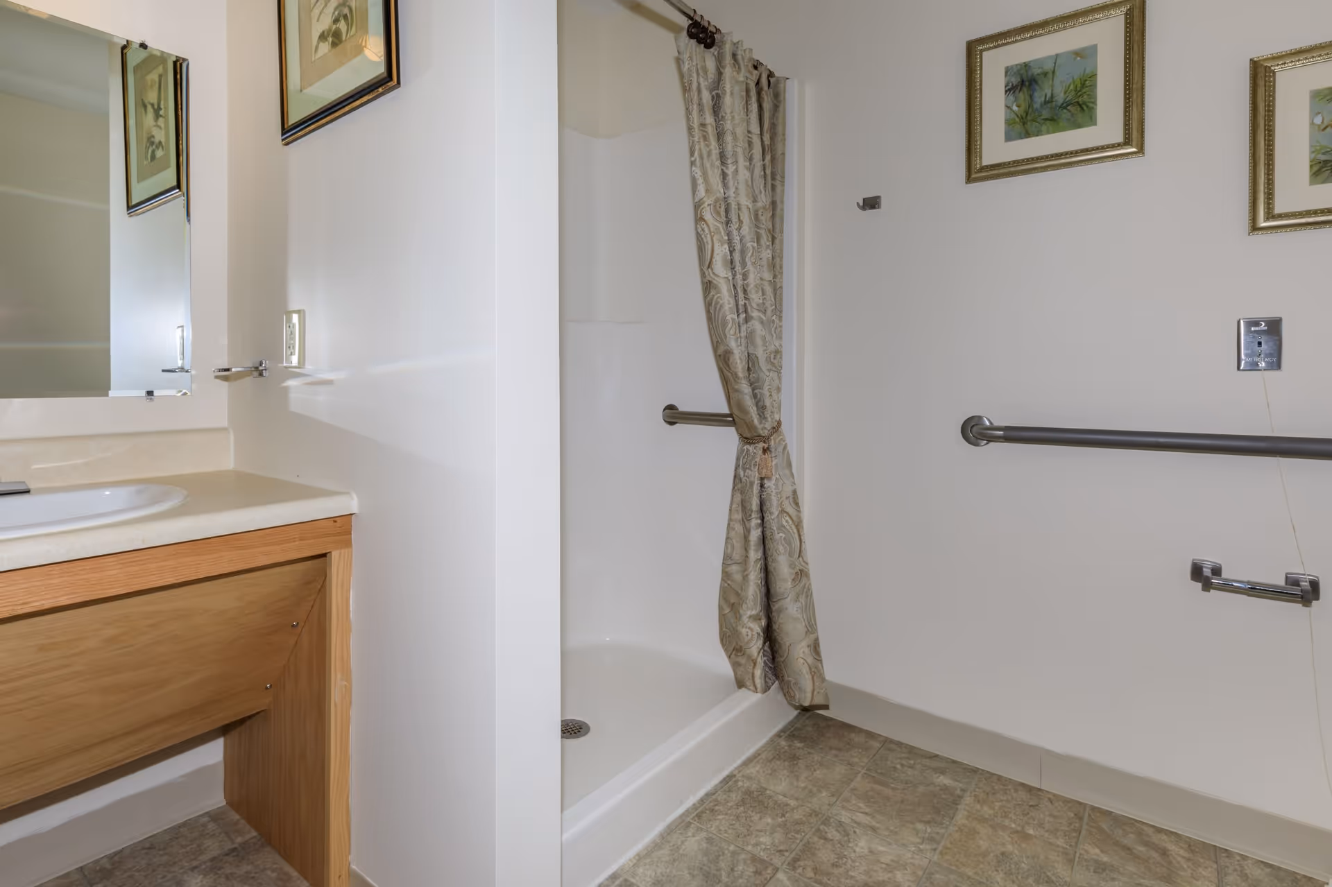A bathroom with a walk-in shower featuring a patterned shower curtain tied back, a grab bar on the wall, and a vanity with a sink and mirror. The walls are white, and there are framed pictures hanging above the grab bar.