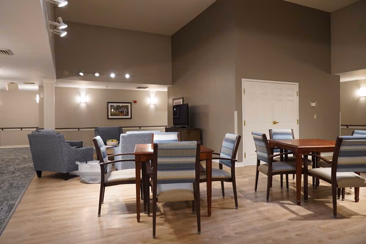 Interior view of a senior living facility common area with wooden tables and chairs arranged for seating. There are upholstered armchairs and a TV on a stand against the wall. The room has beige walls, wood flooring, and soft lighting fixtures.