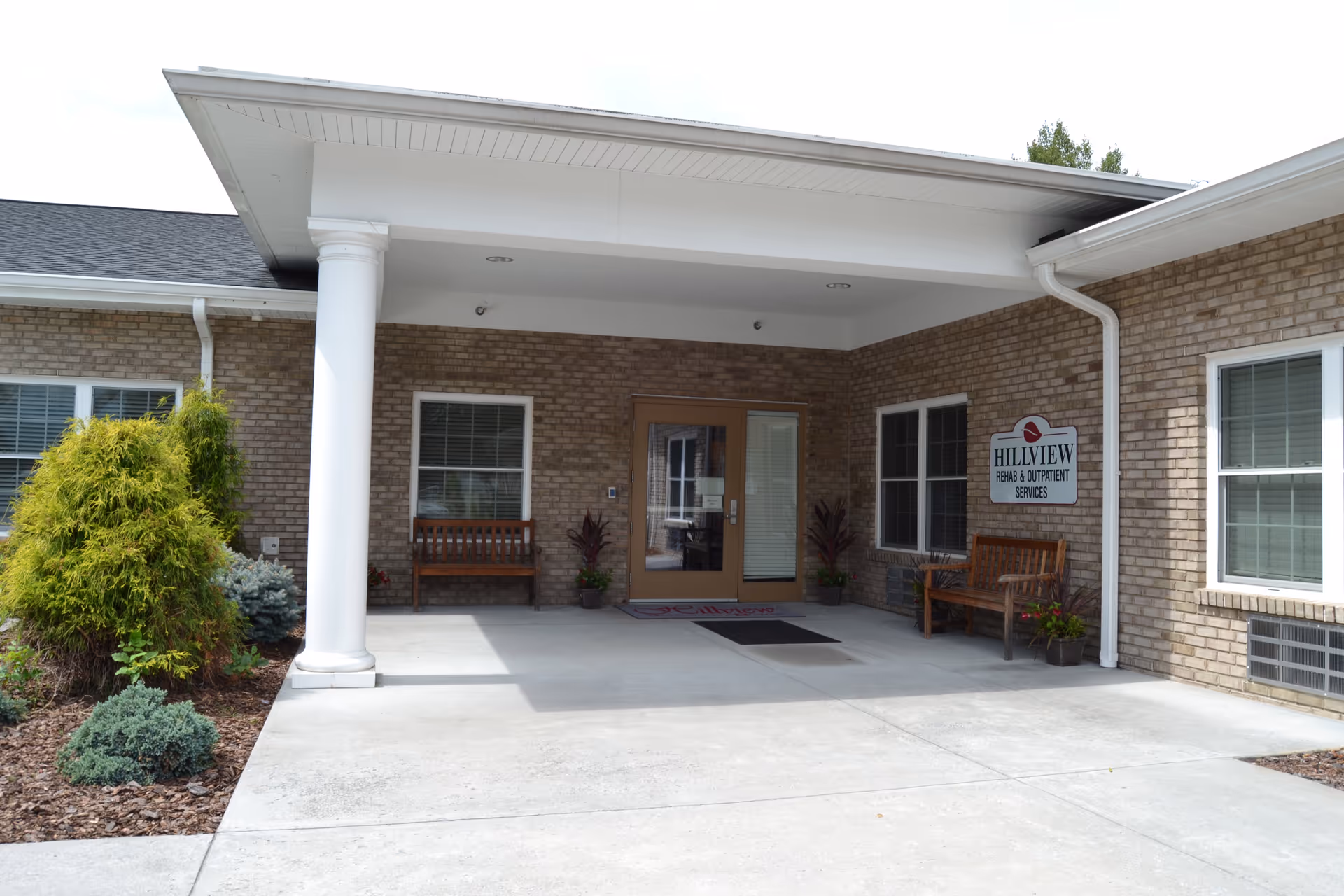 Entrance to Hillview Health and Rehabilitation Center showing a covered porch area with two wooden benches, potted plants, and a sign on the brick wall that reads 'Hillview Rehab & Outpatient Services'. There are windows on either side of the double glass doors, and landscaping with shrubs is visible on the left side.