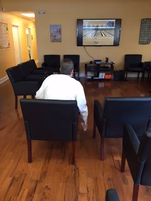 A man sits in a chair in a lounge watching a TV screen showing a bowling alley, with several empty chairs on a hardwood floor.