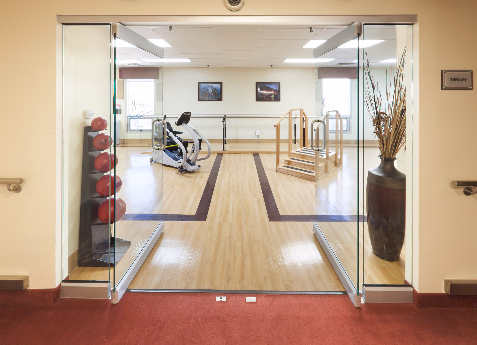 View through glass double doors into a therapy room with exercise equipment including a stationary bike, a set of stairs with handrails for physical therapy, and a rack of red exercise balls. The room has wooden flooring with a dark wood inlay pattern, two windows, and framed pictures on the far wall. A large vase with tall dried plants is placed near the entrance.