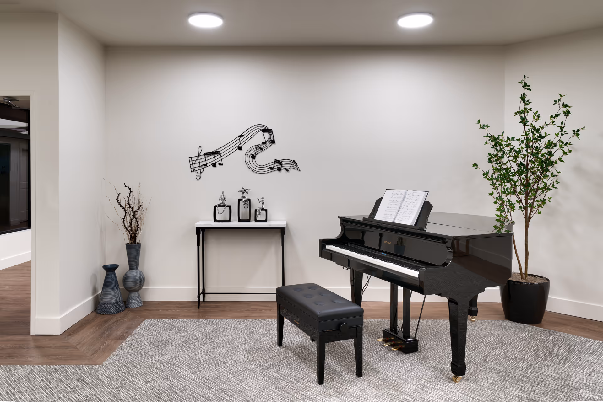 A modern interior room featuring a black grand piano with an open sheet music stand and a matching black piano bench on a gray carpet. To the right of the piano is a tall potted plant with green leaves. On the left side of the room, there is a small table with decorative items and a wall decoration of musical notes above it. The floor is wooden, and the walls are painted white with two ceiling lights illuminating the space.