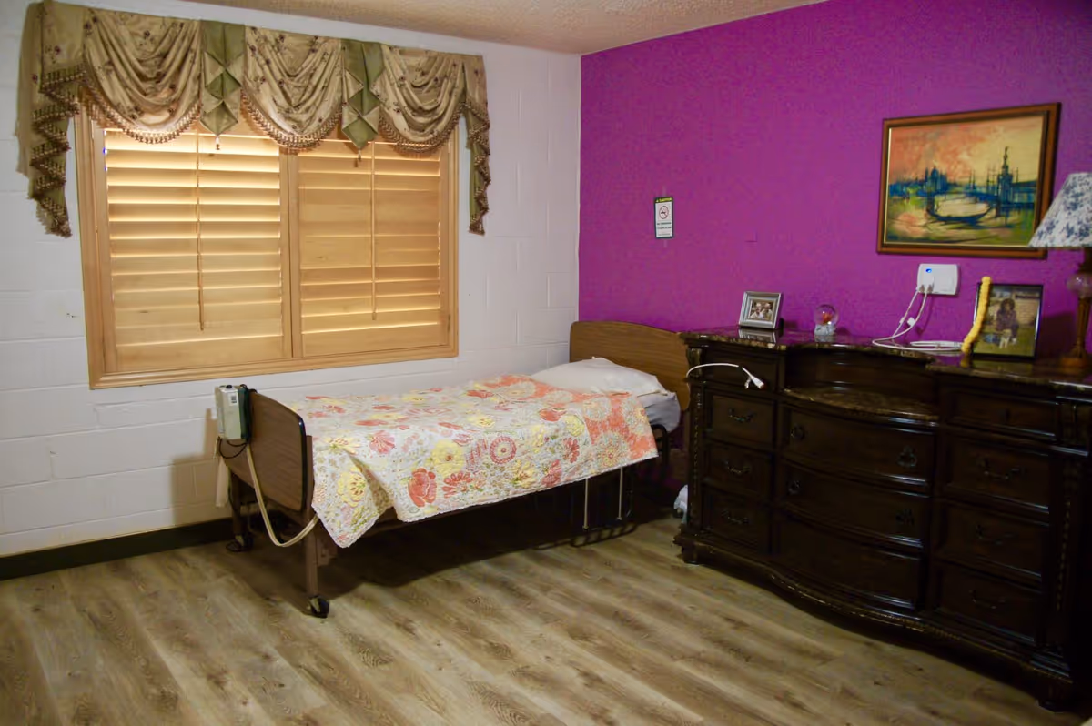 Small bedroom with a single bed covered by a floral quilt, wooden dresser and a purple accent wall with a window.