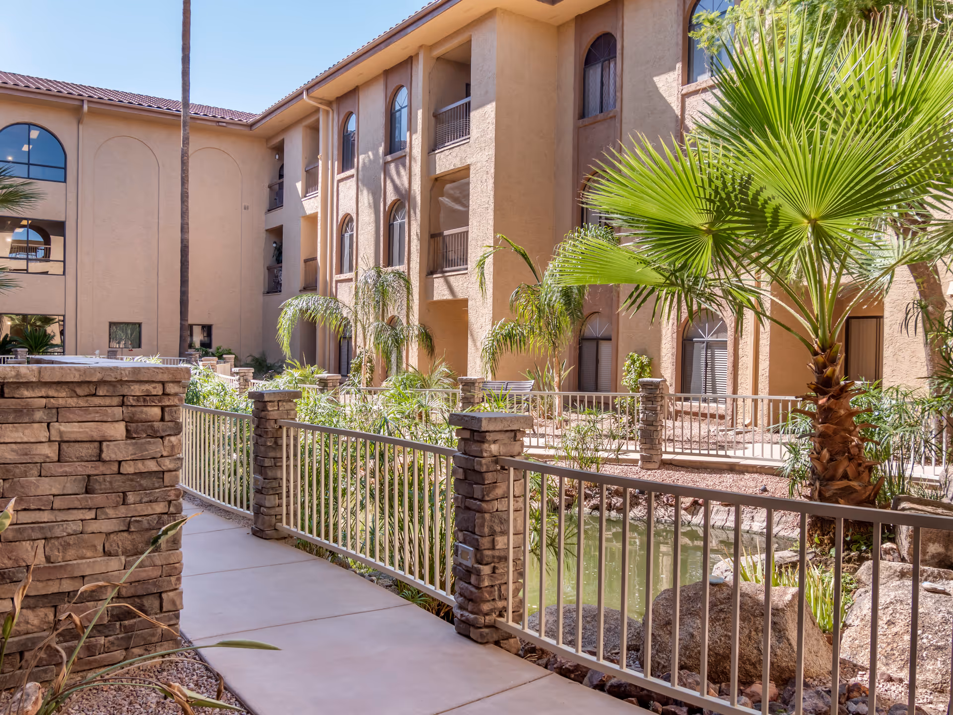 Outdoor courtyard area of a senior living facility with a paved walkway, stone pillars, metal railings, palm trees, and a small pond surrounded by rocks and plants. The beige multi-story building with arched windows is visible in the background under a clear blue sky.