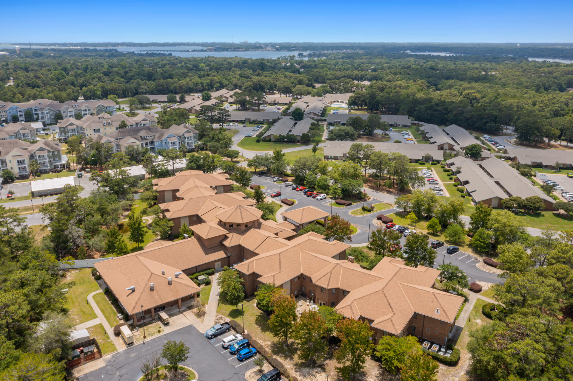 Aerial view of a senior living campus with multiple tan-roofed buildings, parking lots, trees, and a distant body of water.