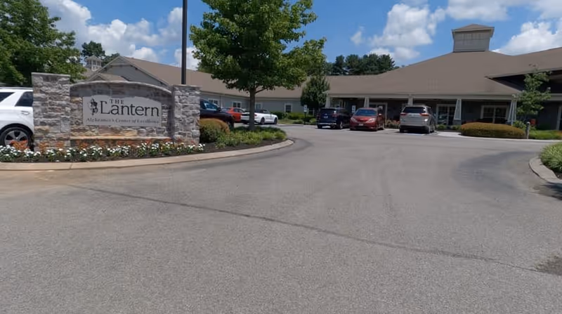 Exterior view of The Lantern Alzheimer's Center of Excellence in Powell, showing the building entrance, parking area with several cars, a stone sign with the facility name, and landscaping with trees and flowers under a partly cloudy sky.