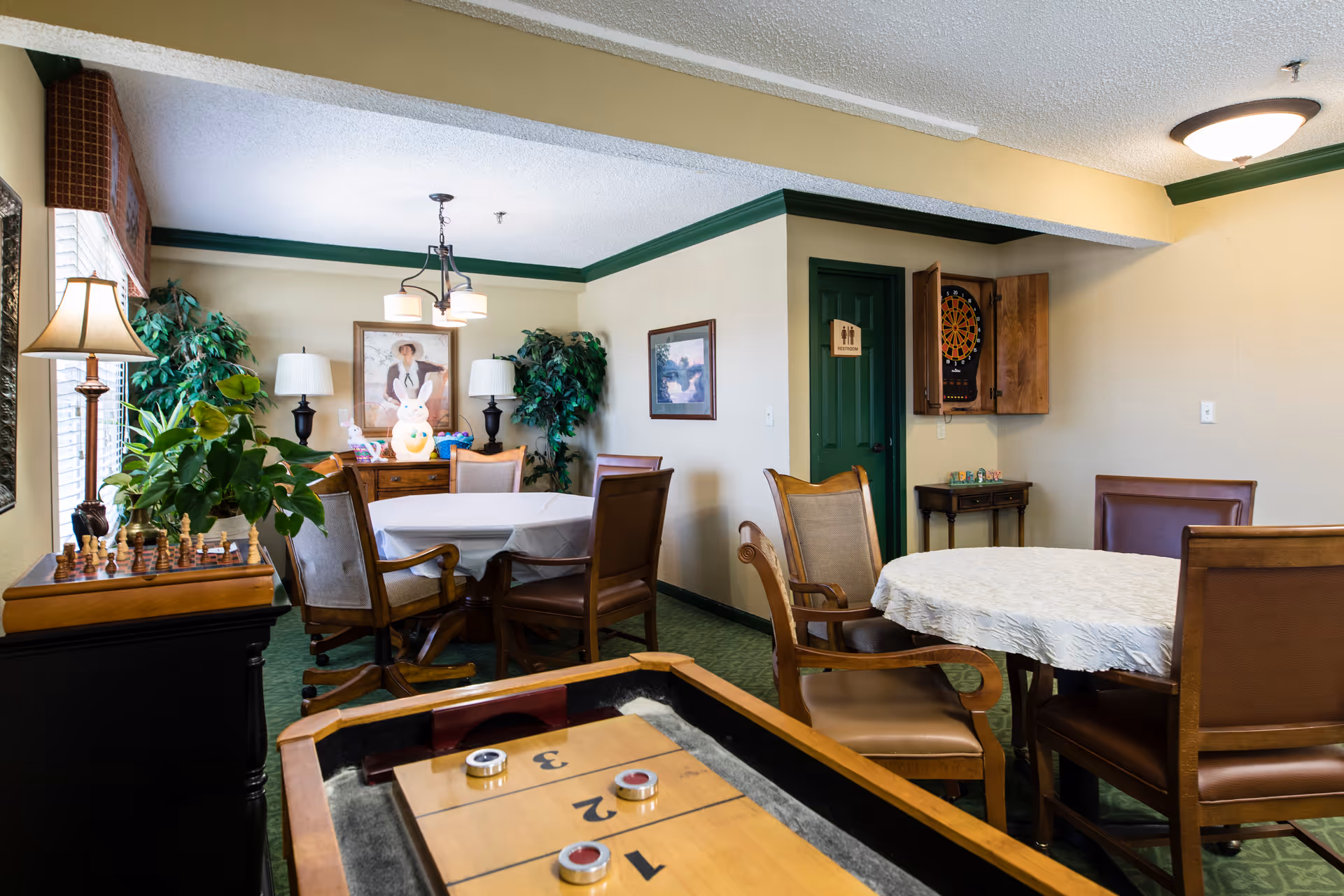 A cozy game room with two round tables covered with white tablecloths surrounded by wooden chairs. There is a shuffleboard table in the foreground, a chess set on a side table to the left, and a dartboard mounted on the wall in a wooden cabinet. The room has green carpet, beige walls with green trim, potted plants, framed artwork, and two table lamps on a wooden sideboard.