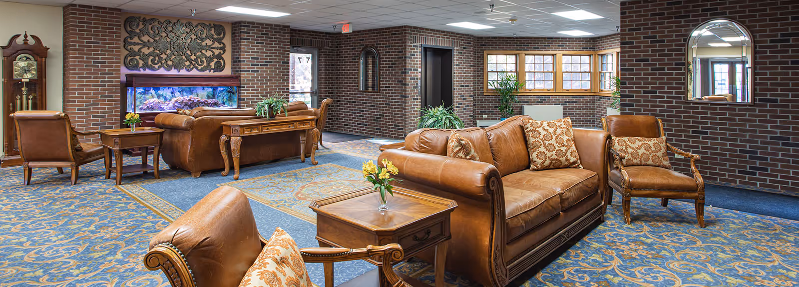 A spacious senior living facility common area with brown leather sofas and armchairs arranged around wooden side tables with small flower vases. The room features a blue patterned carpet, brick walls, a large aquarium built into the wall, a grandfather clock, and a large mirror reflecting the entrance door.