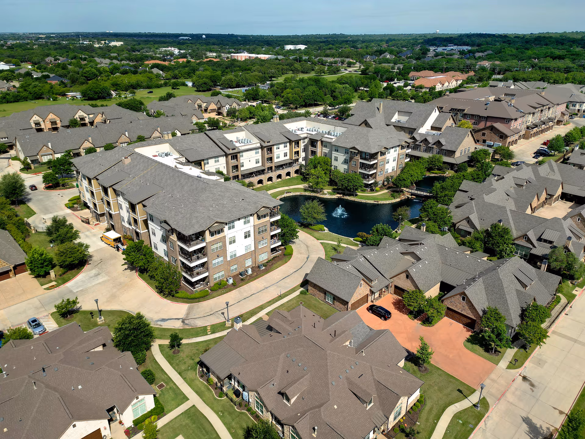 Aerial view of a residential community featuring multiple buildings with gray roofs surrounding a small pond with a fountain in the center. The area is landscaped with trees and pathways, and there are roads and parking areas visible around the buildings.