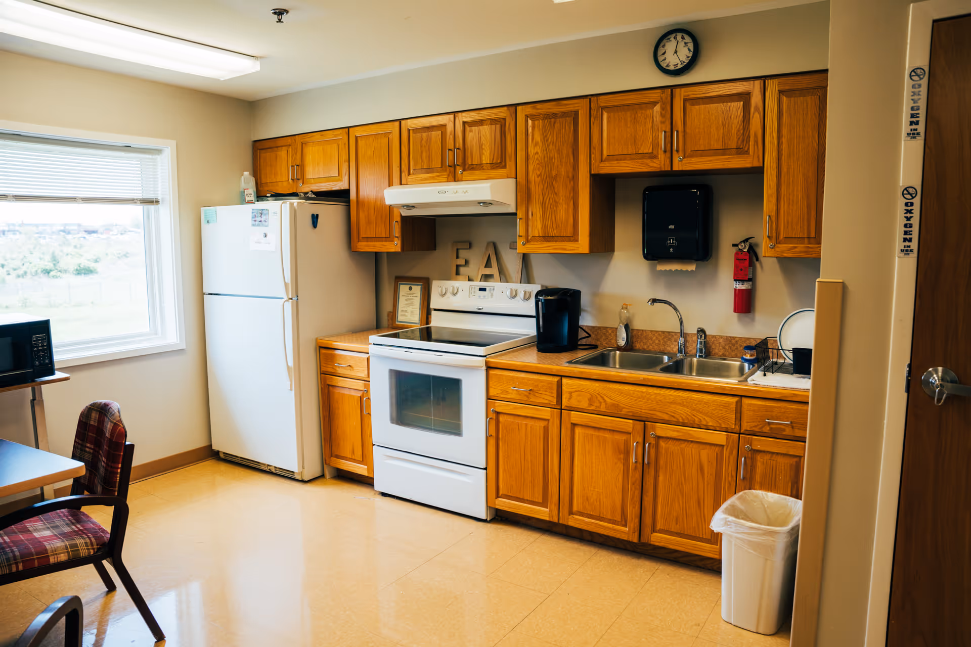 A kitchen area with wooden cabinets, a white refrigerator, a white stove with an oven, a double sink, a microwave on a small table, a clock on the wall, a fire extinguisher, and a trash can. There is a window letting in natural light and a chair with a plaid cushion near a table.
