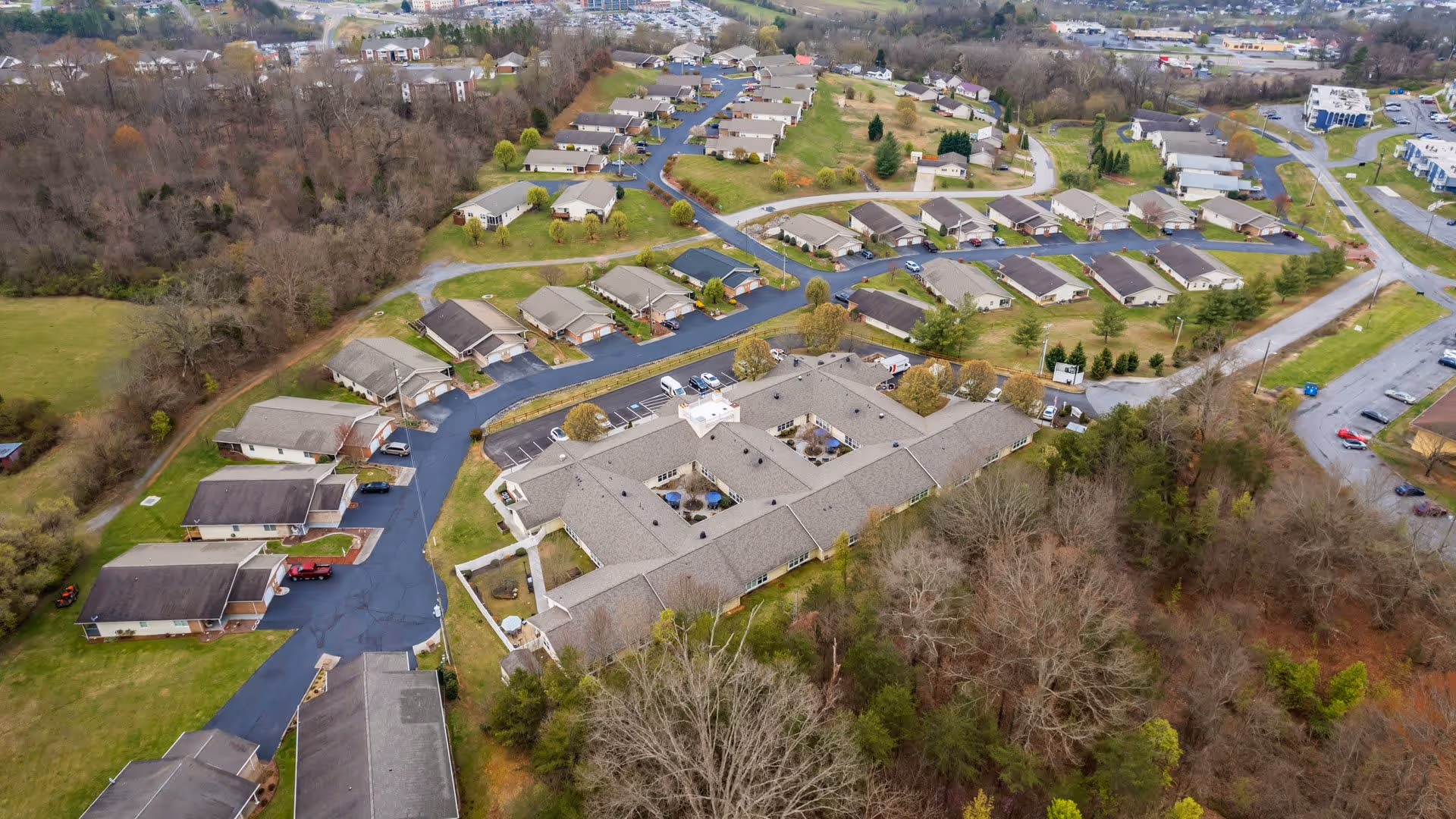 Aerial view of American House Lebanon senior living facility showing a large central building surrounded by multiple smaller residential units, paved roads, parking areas, and trees with a mix of bare and green foliage.