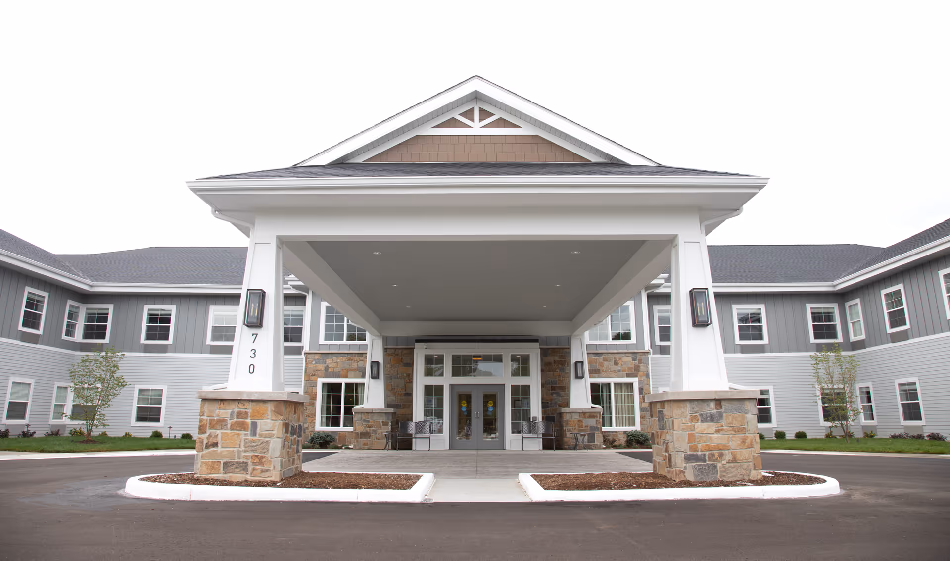 Front exterior view of a senior living facility with a covered entrance supported by white pillars with stone bases, two-story building with gray siding and multiple windows, and a paved driveway.
