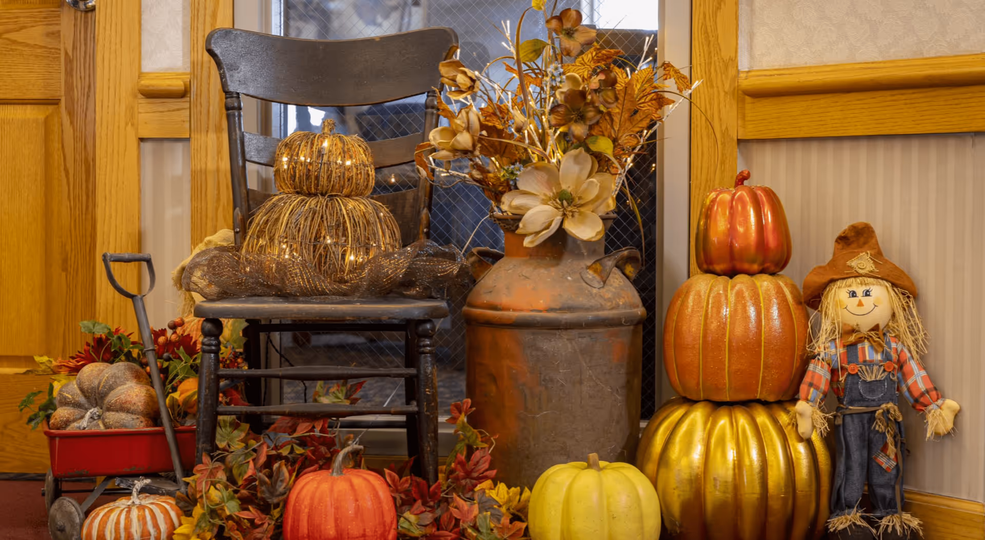Autumn-themed indoor display with pumpkins, gourds, a scarecrow doll, a wooden chair and a vase of dried flowers.