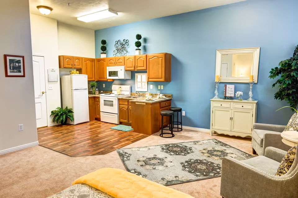 A cozy living space featuring a small kitchen with wooden cabinets, white appliances including a refrigerator, stove, and microwave. The kitchen area has wood flooring, while the rest of the room is carpeted. Adjacent to the kitchen is a small dining counter with two black stools. The living area includes a patterned rug, a beige armchair, a small white cabinet with a mirror above it, decorative candles, and a potted plant. The walls are painted light blue and white, creating a warm and inviting atmosphere.