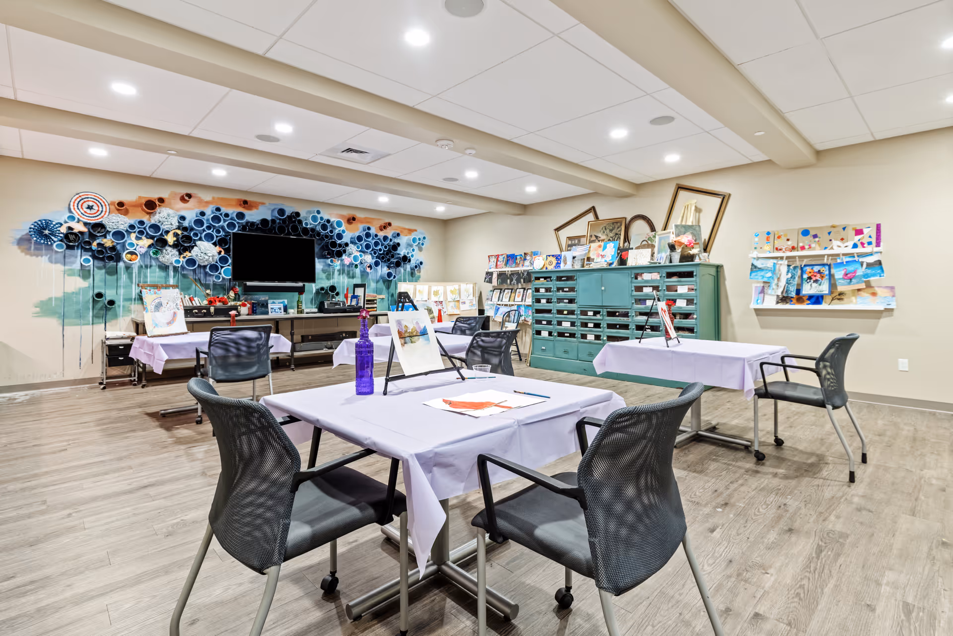 A bright, spacious activity room with tables covered in light purple tablecloths, black mesh chairs, and various art supplies on the tables. The walls display colorful artwork and crafts, with a large decorative wall installation behind a flat-screen TV. The room has wood flooring and recessed ceiling lights.