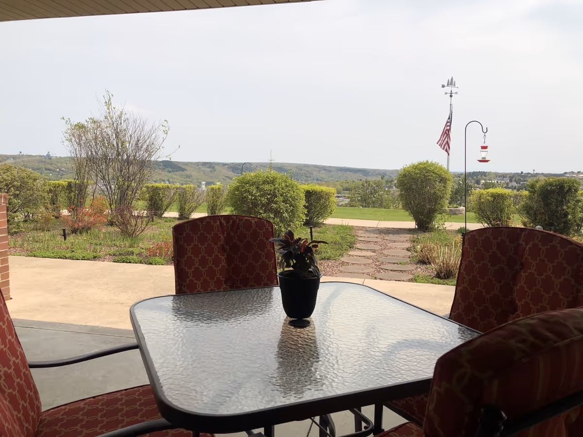 Covered patio with a glass table and red-patterned chairs overlooking a landscaped yard and distant hills.