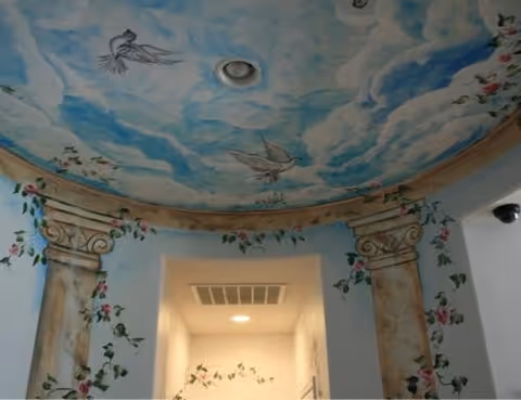 Decorative painted ceiling with clouds and birds, flanked by ornate floral columns inside a facility.