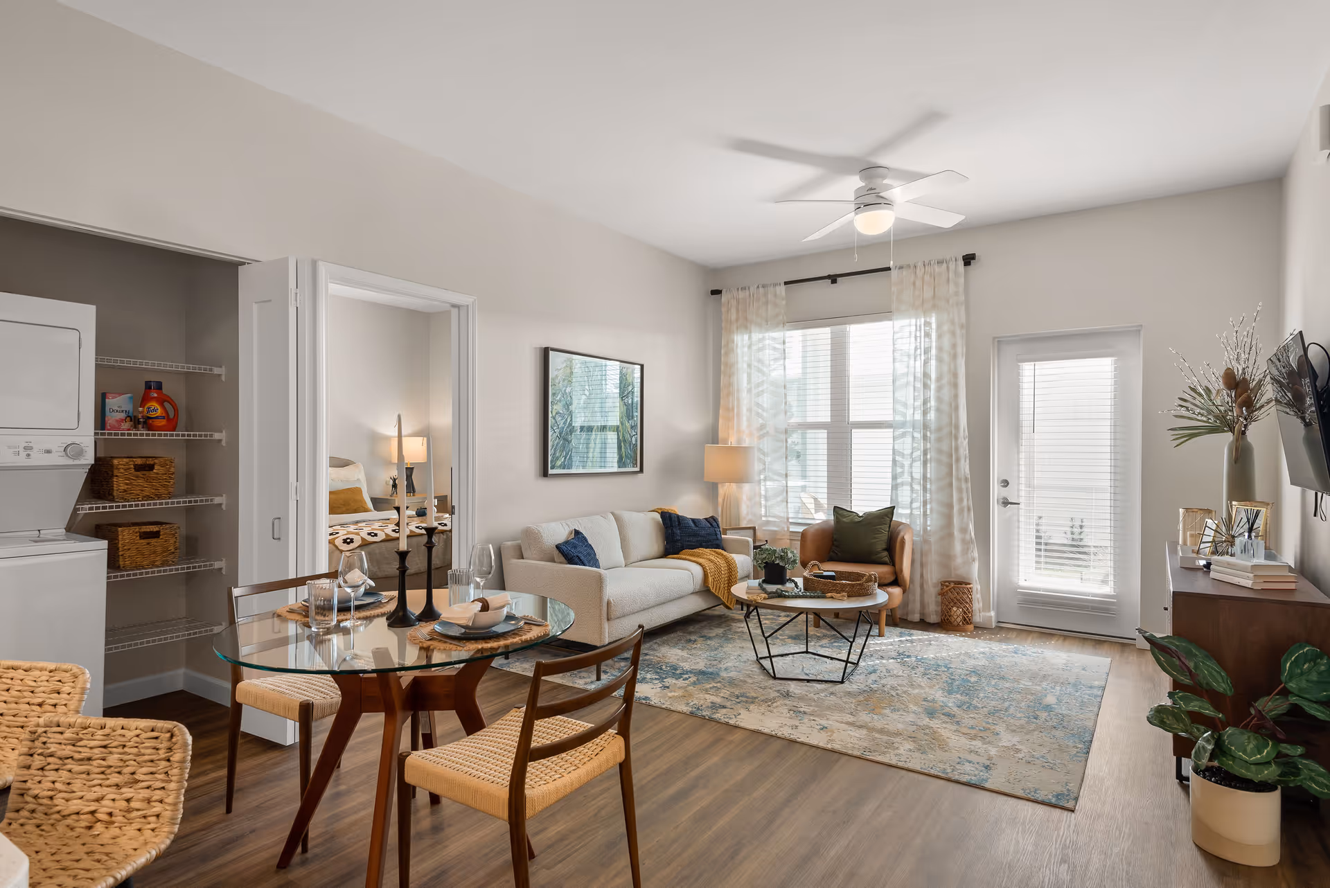 A bright and modern living room with a beige sofa, a brown armchair, a round coffee table on a patterned rug, and a glass dining table with four chairs. There is a ceiling fan with a light, a large window with sheer curtains, a door with blinds, and a wall-mounted TV. To the left, there is a laundry area with a stacked washer and dryer and shelves with laundry supplies. A bedroom with a bed and nightstand is visible through an open doorway.