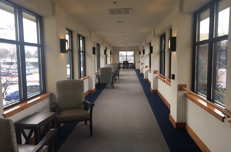 A long, carpeted hallway with large windows on both sides letting in natural light. Several upholstered chairs and small tables are arranged along the left side near the windows. Handrails run along both walls, and wall-mounted lights provide additional illumination. At the far end of the hallway, a table with chairs is visible near a large window.