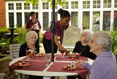 Three elderly women seated around a round outdoor table with red placemats, enjoying fruit plates and drinks, while a caregiver in a purple uniform serves them. The setting is a garden patio with greenery and large windows in the background.