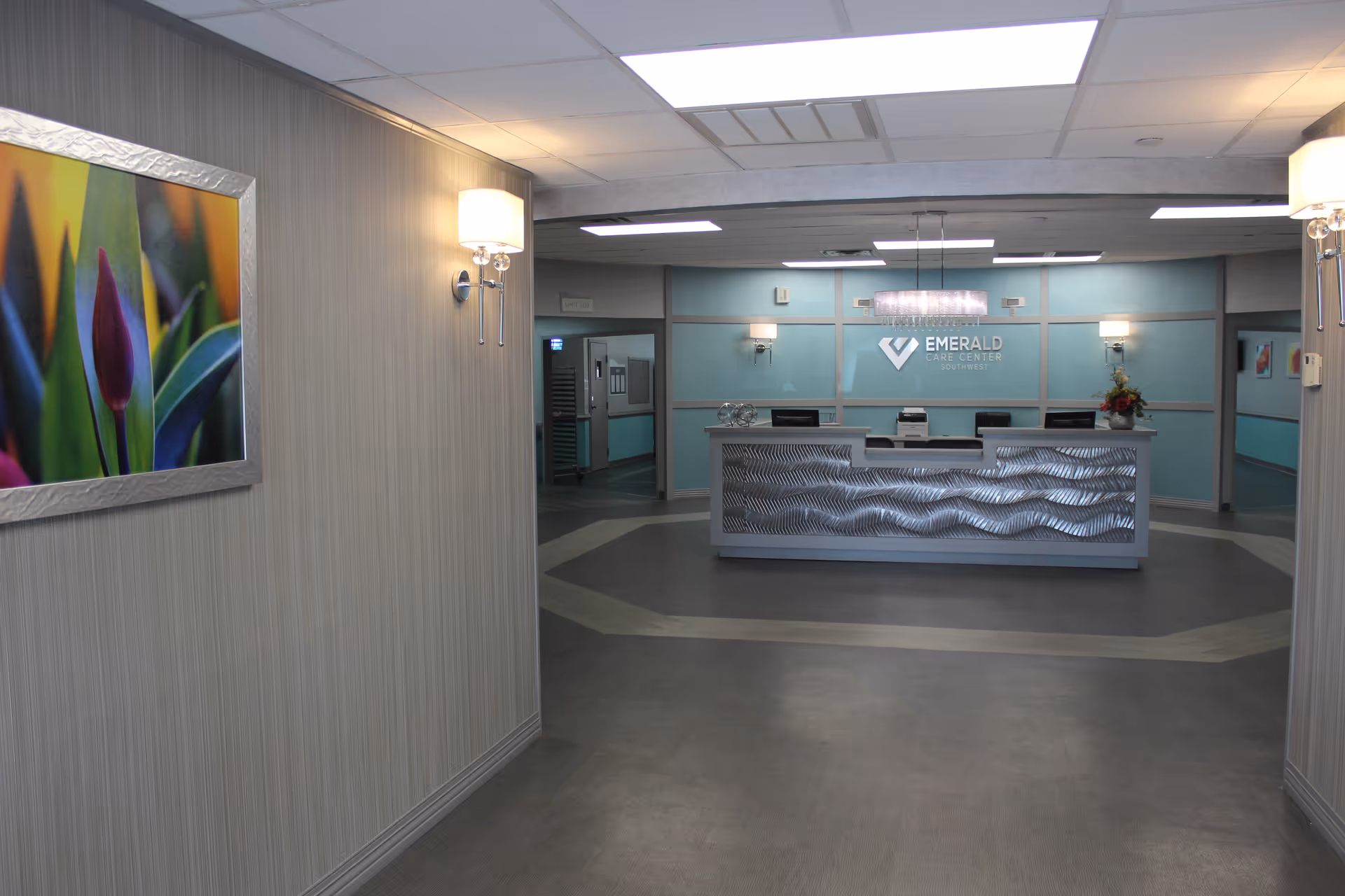 Interior view of the reception area at Emerald Care Center Southwest, featuring a modern front desk with a textured metallic front panel, soft wall lighting, a colorful framed flower photograph on the left wall, and a light blue accent wall behind the desk with the facility's logo.