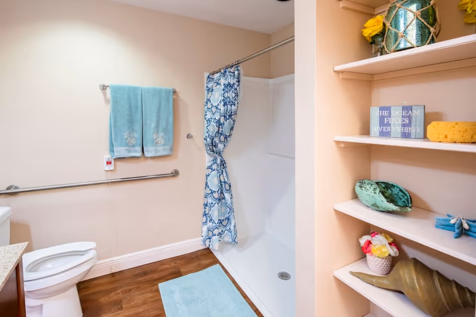 A clean bathroom with a white toilet, a shower area with a blue and white patterned curtain, two blue towels hanging on a wall-mounted towel rack, and a grab bar for support. There is a light blue bath mat on the wooden floor. On the right side, there are built-in shelves decorated with various items including a green vase, a yellow sponge, a sign that reads 'THE OCEAN FIXES EVERYTHING,' and decorative seashells and flowers.