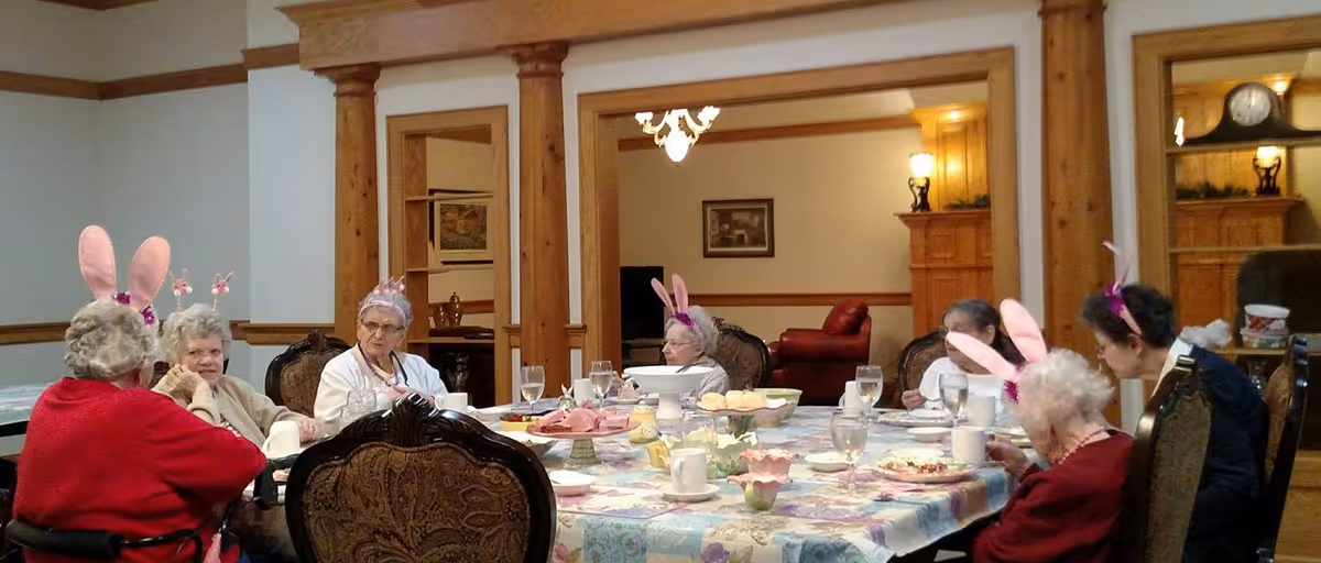 A group of elderly women sitting around a dining table in a retirement facility, some wearing pink bunny ears headbands. The table is set with plates, cups, glasses of water, and various food items. The room has wooden pillars and furniture, with a cozy and warm atmosphere.