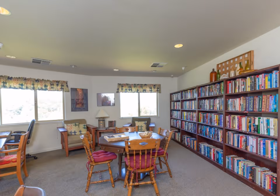 A cozy senior living facility room with a round wooden table surrounded by four wooden chairs with red cushions. There are two armchairs with patterned pillows near two windows with floral valances. A large bookshelf filled with books lines one wall, and there are framed pictures and a lamp on a small table between the armchairs. The room is well-lit with ceiling lights and natural light from the windows.