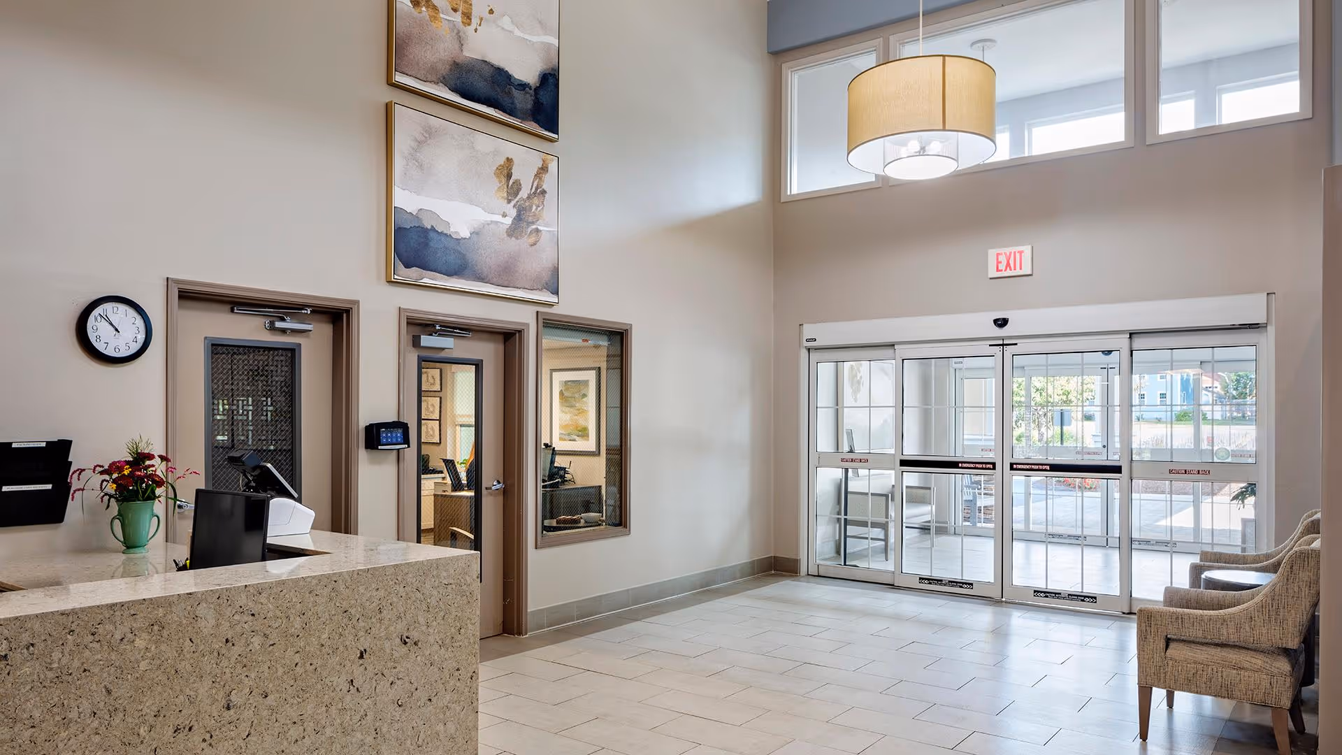 Bright and spacious lobby area of a senior living facility with a reception desk on the left, two closed doors behind the desk, a clock on the wall, two abstract paintings above the doors, and a large glass automatic sliding entrance door on the right. There are two upholstered chairs near the entrance and a hanging ceiling light fixture.