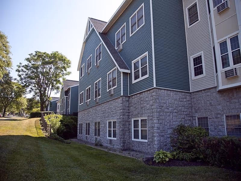 Three-story senior living building with green siding and a stone foundation next to a manicured lawn and trees.