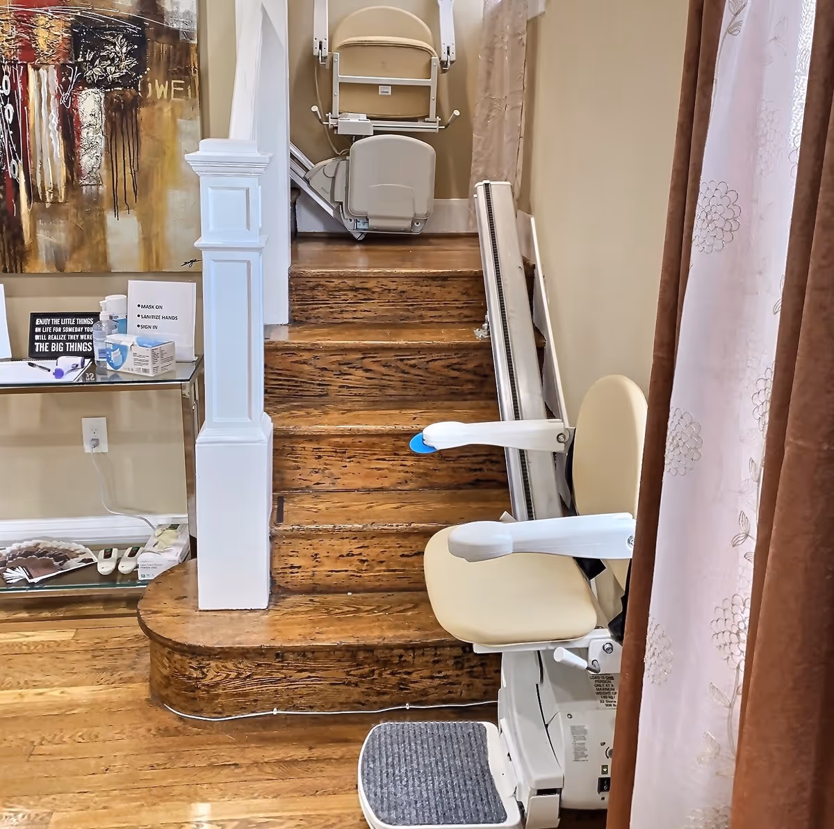 Wooden staircase with a beige stairlift installed on the right side. The stairlift has a padded seat and armrests. To the left of the stairs is a small glass table with various items including a sign, sanitizer, and a box. A colorful abstract painting hangs on the wall above the table. Beige walls and a curtain with floral patterns are visible on the right side.