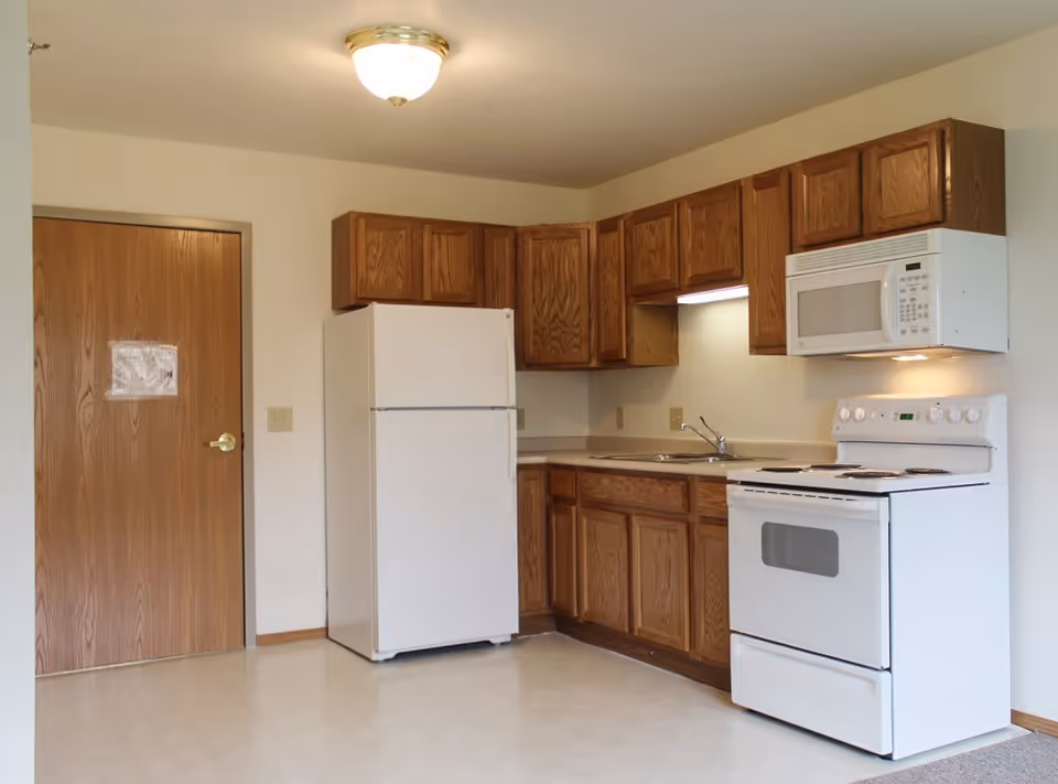A kitchen area with wooden cabinets, a white refrigerator, a white electric stove with oven, and a white microwave mounted above the stove. There is a sink with a faucet under the cabinets. The floor is light-colored, and there is a wooden door to the left side of the kitchen.