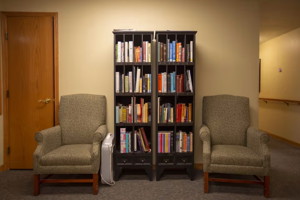 Two patterned armchairs with wooden legs are positioned on either side of two tall black bookshelves filled with books. A wooden door is visible on the left side, and a hallway with handrails is visible on the right side. The walls are painted beige and the floor is carpeted.