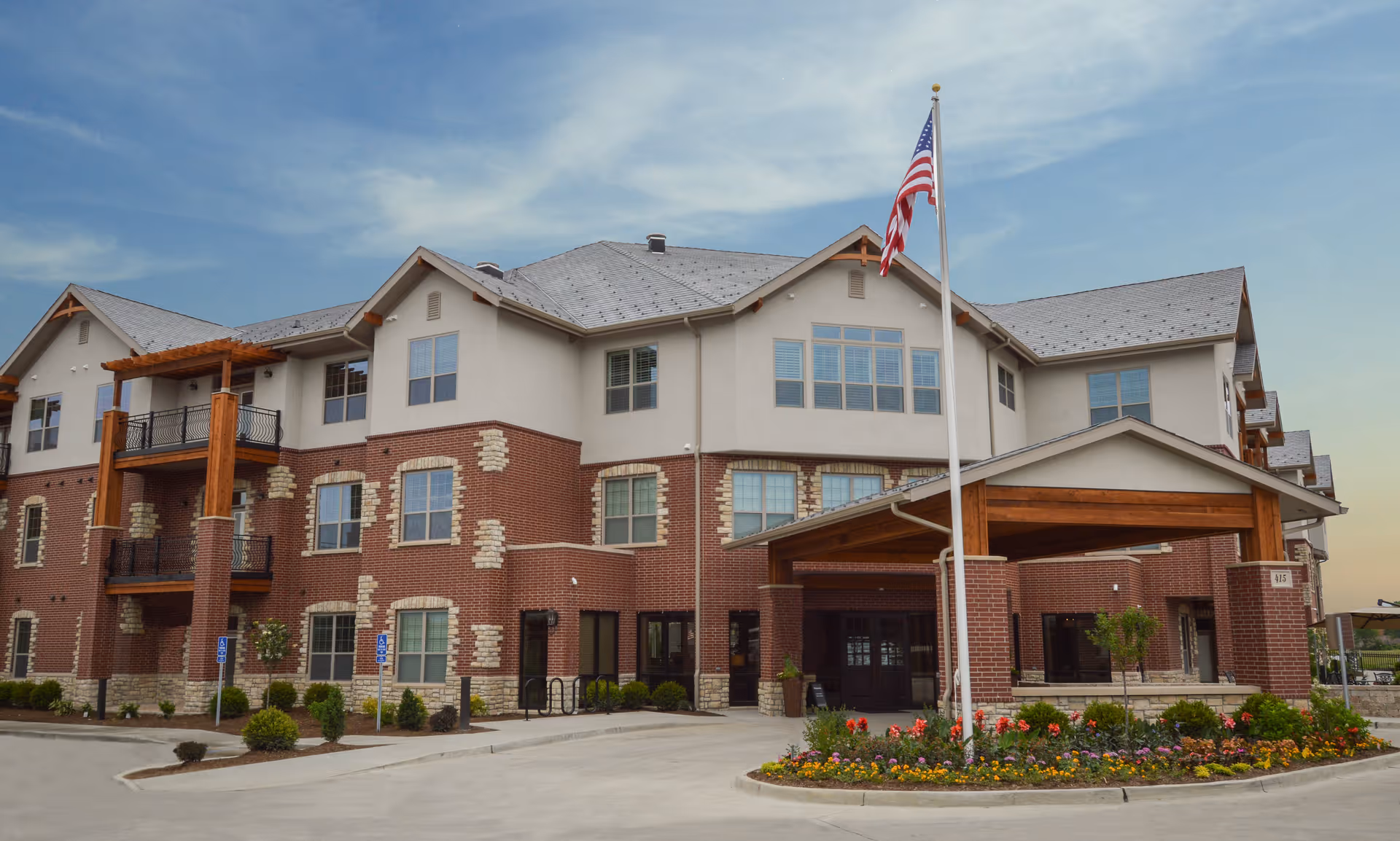 Exterior view of a three-story senior living facility building with a combination of brick and light-colored siding. The entrance features a covered drop-off area with wooden beams, and an American flag is prominently displayed on a flagpole in front of a landscaped flower bed. The sky is partly cloudy with a soft evening light.