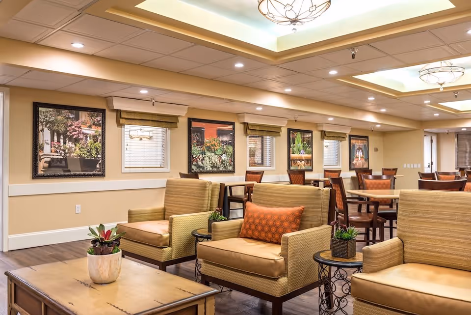 A well-lit common area in a senior living facility featuring comfortable armchairs with cushions, wooden tables with small potted plants, and several dining tables with chairs in the background. The walls are decorated with framed pictures of plants and flowers, and the ceiling has recessed lighting and decorative light fixtures.