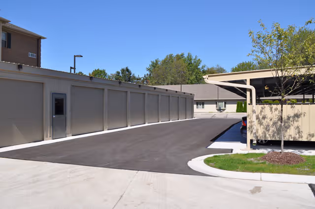 Paved driveway between a row of storage/garage units and a covered carport beside the facility with a building and trees in the background.