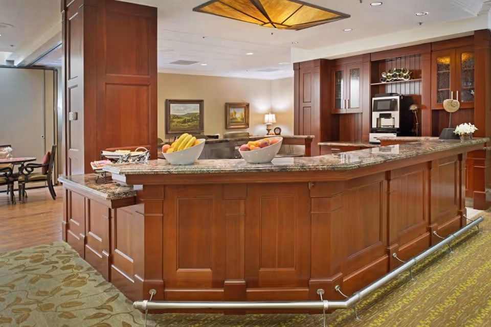 Wood-paneled communal service counter with a granite countertop, bowls of fruit, and a coffee station in a senior living common area.